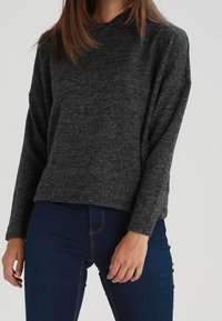 Ciemnoszary sweter typu pullover o luźnym kroju, długich rękawach i miękkiej fakturze. W zestawieniu z niebieskimi dżinsami z kontrastowymi przeszyciami.