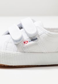 Sneaker bianca in tela con doppio strap in velcro, lato testurizzato e suola in gomma con un motivo di aderenza; logo brandizzato in rosso e blu.