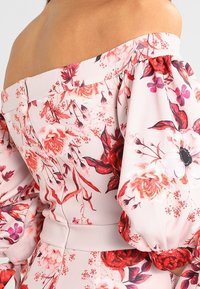 Kwiatowa bluzka bez ramiączek z wyrazistymi czerwonymi i różowymi wzorami na jasnoróżowej tkaninie, z marszczonymi bufkami i dopasowanym w pasie.