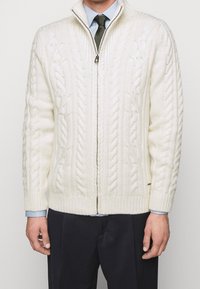 Krämfärgad kabelstickad cardigan med hög krage och frontdragkedja. Ribbad manschett och nederkant. Buren över en ljusblå skjorta och slips.