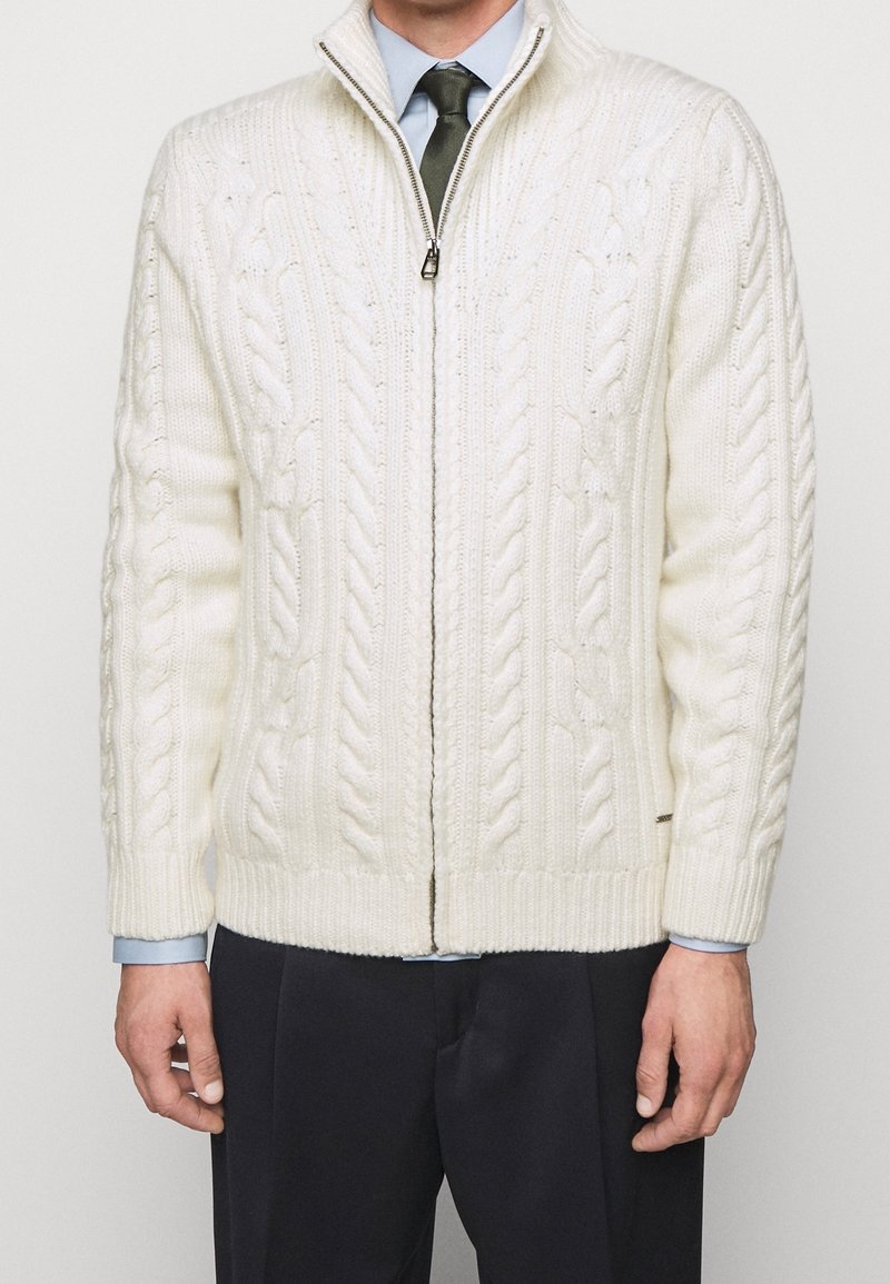 Krämfärgad kabelstickad cardigan med hög krage och frontdragkedja. Ribbad manschett och nederkant. Buren över en ljusblå skjorta och slips.