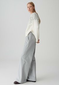 Maglione leggero crema con un giromanica grigio a motivo, abbinato a pantaloni larghi a righe grigie. Il modello è in posizione laterale, mettendo in evidenza i dettagli dell'outfit.