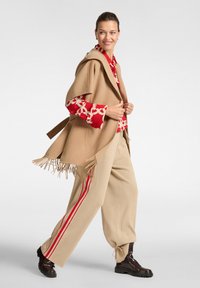 Poncho beige con orlo frangiato, indossato sopra una blusa a righe rosse e bianche. Pantaloni beige con strisce laterali rosse e stivaletti marroni alla caviglia.