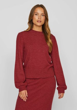 Femme aux longs cheveux châtain clair portant un pull en maille rouge avec des manches bouffantes et une jupe côtelée assortie sur un fond gris uni.