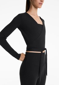 Black long-sleeve wrap top with a v-neckline, featuring a tied waist. The fabric has a ribbed texture, paired with high-waisted leggings.