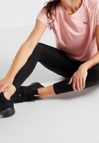 Camiseta deportiva rosa de Puma y leggings negros; zapatillas negras con diseño texturizado y logo; modelo sentado, estirando la pierna hacia adelante.