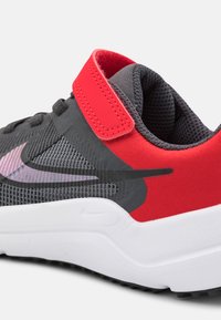 Scarpa sportiva grigia con accenti rossi, tomaia in mesh e cinturino in velcro. Presenta una suola bianca e un logo riflettente sul lato.