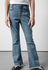 Osoba ubrana w niebieskie dżinsy z wysokim stanem, z guzikami, postrzępionymi brzegami i czarnymi butami na slip-on z okrągłymi wycięciami, stojąca na szarym podłożu.