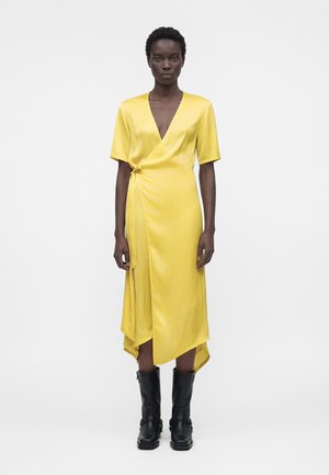 Frau steht frontal, trägt ein gelbes Wickelkleid mit kurzen Ärmeln und schwarze Stiefel bis zur Mitte der Wade vor weißem Hintergrund.