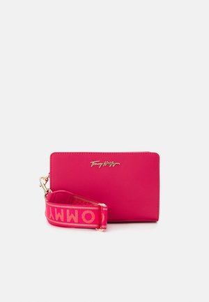 Jasnoróżowy, prostokątny portfel z złotym logo "Tommy Hilfiger" i odpinanym paskiem z pogrubionymi różowymi literami na czerwonym tle.