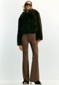 Donna che indossa un giubbotto in pelliccia sintetica oversize marrone scuro, pantaloni a zampa d'elefante marroni aderenti, tacchi aperti pelosi e occhiali da sole grandi su uno sfondo bianco.