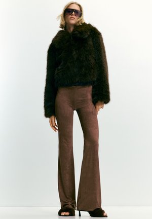 Ženska oblečena v prenajeto temno rjavo umetno krzno jakno, rjave oprijete široke hlače, puhaste sandale z odprtim prstom in velike sončne očala na belem ozadju.