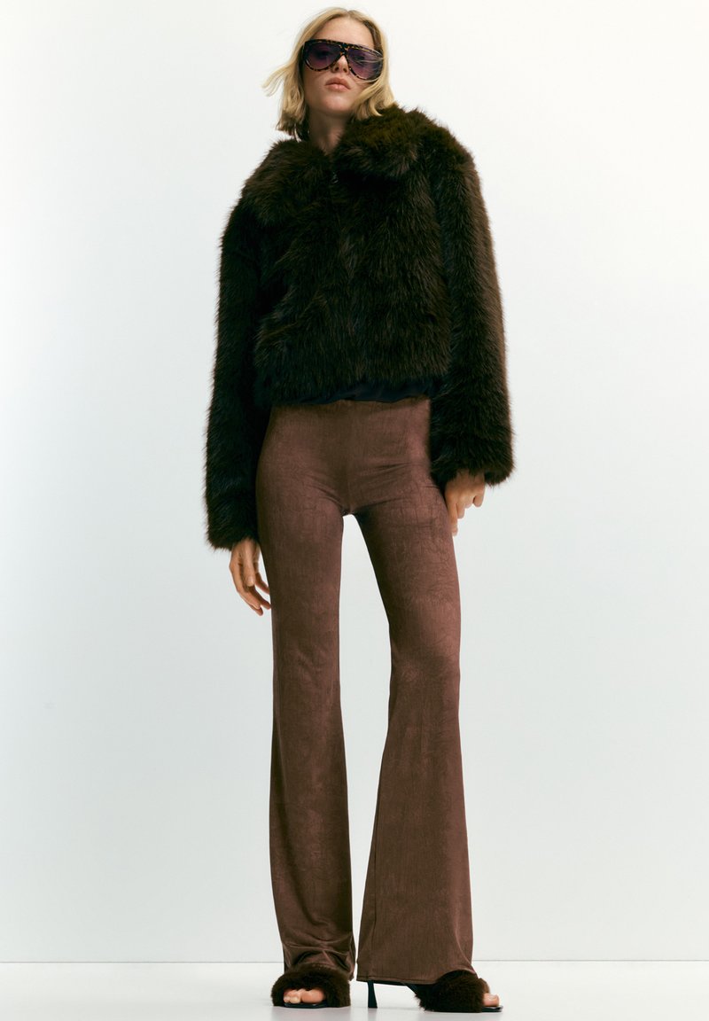 Donna che indossa un giubbotto in pelliccia sintetica oversize marrone scuro, pantaloni a zampa d'elefante marroni aderenti, tacchi aperti pelosi e occhiali da sole grandi su uno sfondo bianco.