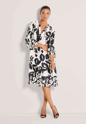 Schwarz-weißes Kleid mit Blumenmuster, 3/4-Ärmeln, einem V-Ausschnitt und einer legeren Passform, kombiniert mit schwarzen offenen Schuhen mit Absatz.