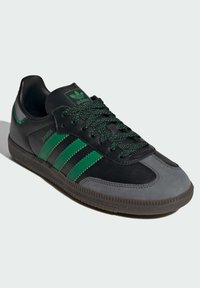 Black leather sneakers with green accents, featuring a gray toe cap and three striped design. Textured rubber sole with textured laces.