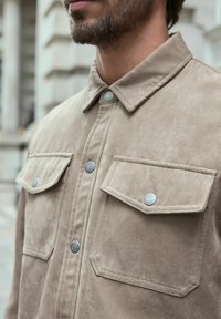 Cămașă din suede bej cu guler ascuțit, două buzunare frontale și închidere cu capse. Textură moale cu un finisaj neted și detalii discrete de cusătură.
