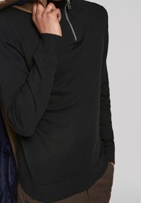 Pullover nero a maglia con colletto alto e dettagli in zip argentata. Polsini e orlo a costine; tessuto leggero. Indossato sotto una giacca marrone.