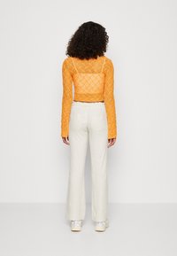 Une femme aux cheveux bouclés porte un top en dentelle orange à manches longues, un pantalon blanc taille haute et des baskets blanches, se tenant devant un fond uni.