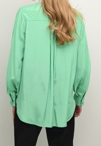 Blusa in satin verde menta con maniche lunghe a sbuffo, orlo arrotondato e polsini con bottoni. Texture liscia, design minimalista e vestibilità rilassata.