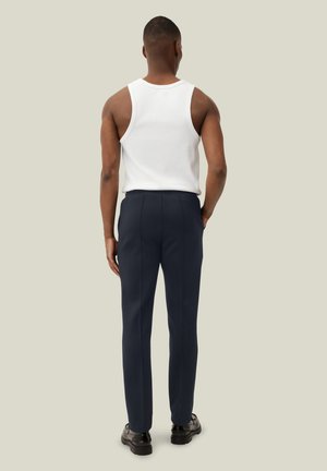 Homme debout de dos portant une chemise blanche sans manches, un pantalon bleu marine sur mesure et des chaussures habillées noires sur un fond uni.