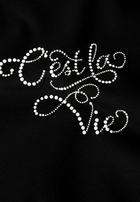 Melnas audums ar baltām pērlītēm, kas veido tekstu "C'est la Vie" eleganti izliektā dizainā, ar dažāda izmēra pērlītēm, kas uzsver struktūru.