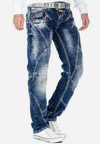 Jeans in denim di colore blu scuro con finitura rovinata. Presentano intricate cuciture e due tasche frontali. Indossati con sneakers bianche.