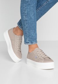 Beige kanvas sneakers med en tjock vit plattformssula, rund tå och metallögon för snörningen. Texturerad ovandel med sidostripade detaljer.