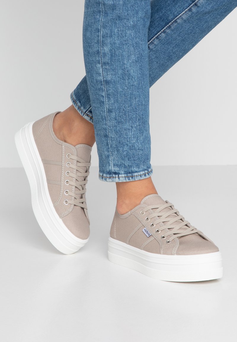 Beige kanvas sneakers med en tjock vit plattformssula, rund tå och metallögon för snörningen. Texturerad ovandel med sidostripade detaljer.