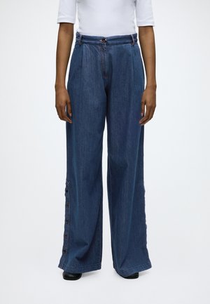 Persoon draagt een blauwe denim broek met hoge taille en wijde pijpen, met knoopdetails langs de buitenkant van één pijp.