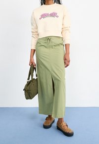 Persona che indossa una felpa color crema con logo rosa, una gonna verde oliva con spacco anteriore, scarpe con stampa leopardata e tiene una borsa verde oliva.