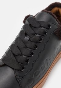 Svarta lädersneakers med texturerad yta, platta svarta snören, foder i fuskpäls och gummisulor i brunt. Har ett präglat logotyp på sidan.