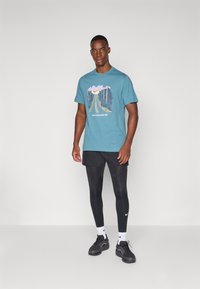 Camiseta de algodón en color verde azulado con un gráfico de montaña y texto, combinada con pantalones cortos negros, leggings negros y zapatillas negras. Diseño sencillo, ajuste casual.