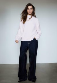 Lys rosa strikket sweater med ribbede manchetter, parret med vidde denim jeans i mørk farve med synlige sømmene. Enkel baggrund.