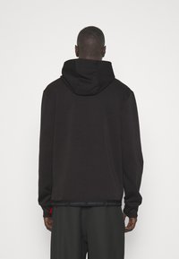 Schwarzer Kapuzenpullover mit langen Ärmeln, der am Saum ein Logo-Band aufweist. Der Stoff wirkt weich mit einer glatten Textur.