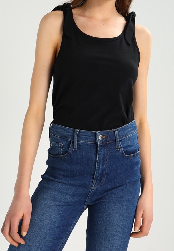 Vrouw in een zwart mouwloos top met gestrikte schouderbanden en hooggesneden blauwe spijkerbroek tegen een effen achtergrond.