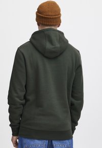 Dark green hoodie with a textured, soft fabric. Features a large hood and ribbed cuffs, paired with a brown knitted beanie.