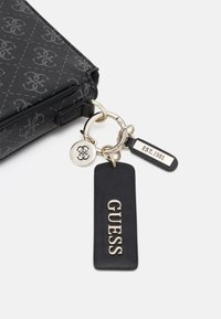 Schwarzes strukturiertes Wallet mit Logo-Muster, silbernem Hardware und Schlüsselanhänger mit einem "GUESS"-Tag und einer "EST. 1981"-Plakette.