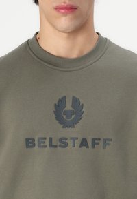 Maslinasto zeleni dukserica s rundim izrezom, s teksturiranim crnim logotipom i nazivom marke "BELSTAFF" ispisanim velikim slovima.