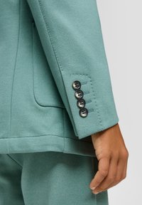 Turkusowy garnitur ze skrojonym krojem i fakturowanym wykończeniem, z mankietem zapinanym na cztery czarne guziki. Widoczna ręka, pokazująca szczegóły.