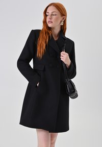 Cappotto in misto lana nero con design doppiopetto, bottoni arrotondati, tasche laterali e una silhouette sartoriale. La texture appare liscia.