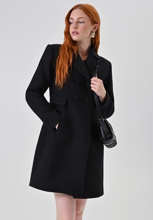 Cappotto in misto lana nero con design doppiopetto, bottoni arrotondati, tasche laterali e una silhouette sartoriale. La texture appare liscia.