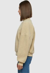 Sweatshirt en polaire beige avec un col haut, des épaules tombantes et des poignets côtelés. Texture douce et coupe décontractée, associé à un jean bleu.