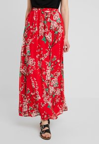 Jupe longue rouge à fleurs avec une coupe ample, présentant des motifs de fleurs blanches et vertes, une taille froncée et un tissu léger et fluide.