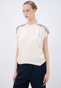 Haut à manches courtes crème avec des accents de perles sur les épaules, en tissu doux, coupe décontractée, associé à un pantalon texturé bleu marine.