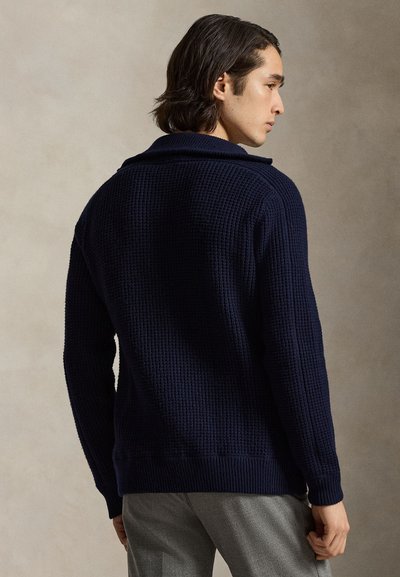 Navy blue knit sweater with a textured pattern, shawl collar, and ribbed cuffs and hem. View from the back, model wearing gray pants.