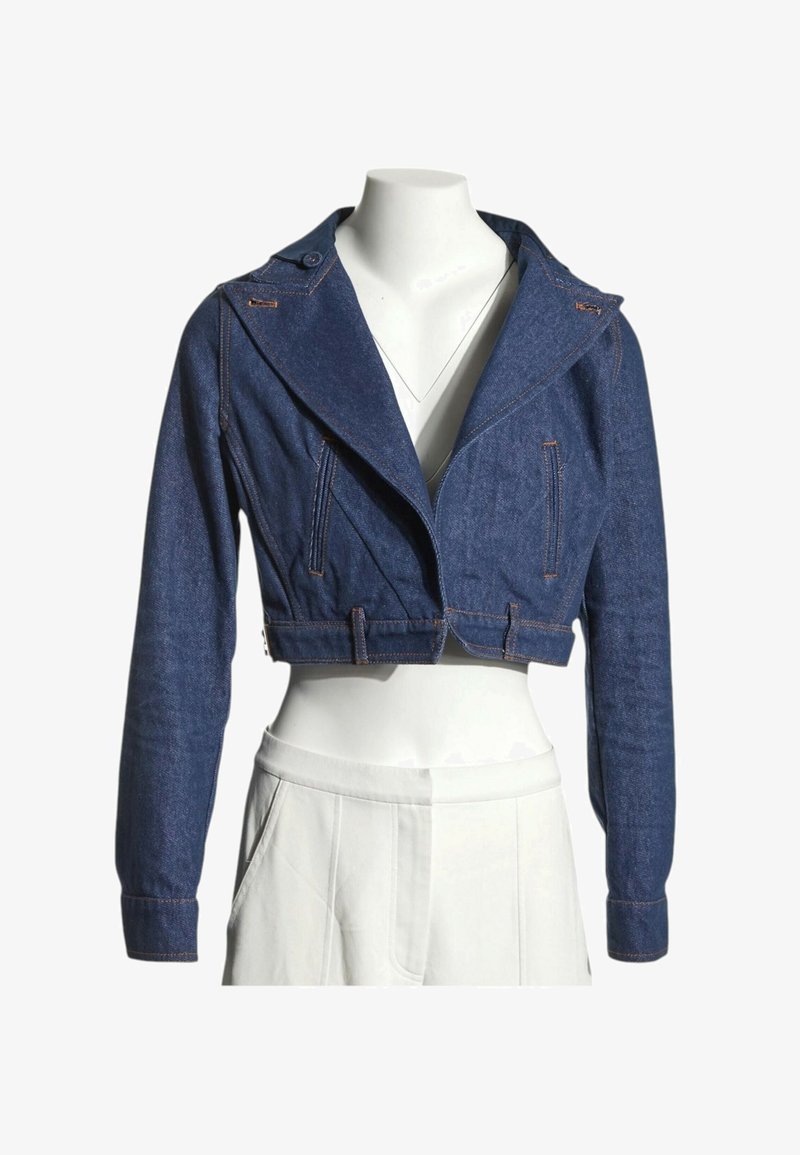 Veste en jean courte bleu foncé avec manches longues, larges revers et détail ceinture, portée sur un mannequin blanc avec un pantalon blanc.