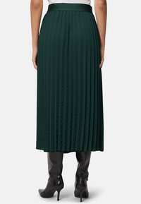 Jupe maxi plissée vert foncé avec une taille lisse et des plis verticaux, associée à des bottes noires hauteur genoux.