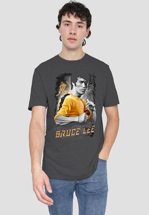 Dunkelgraues T-Shirt mit einem gelben Grafik von Bruce Lee in Aktion, darunter der Text "BRUCE LEE" in Orange. Lässiger Stil.