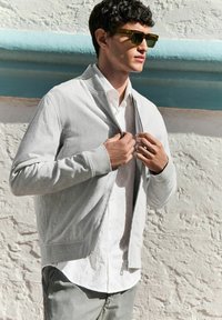 Chaqueta gris con cremallera y acabado texturizado, llevada sobre una camisa blanca con estampado, complementada con gafas de sol verdes y pantalones grises.