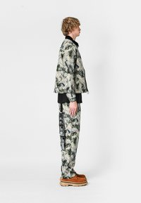 Chaqueta y pantalones con estampado de camuflaje en verde y beige, con detalles en negro. Calzado marrón con una suela gruesa. Tejido texturizado.